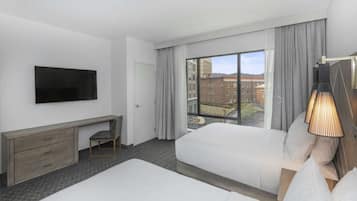 A hotel room with two beds, a TV mounted on the wall, a desk with drawers, a chair, and a large window with curtains.