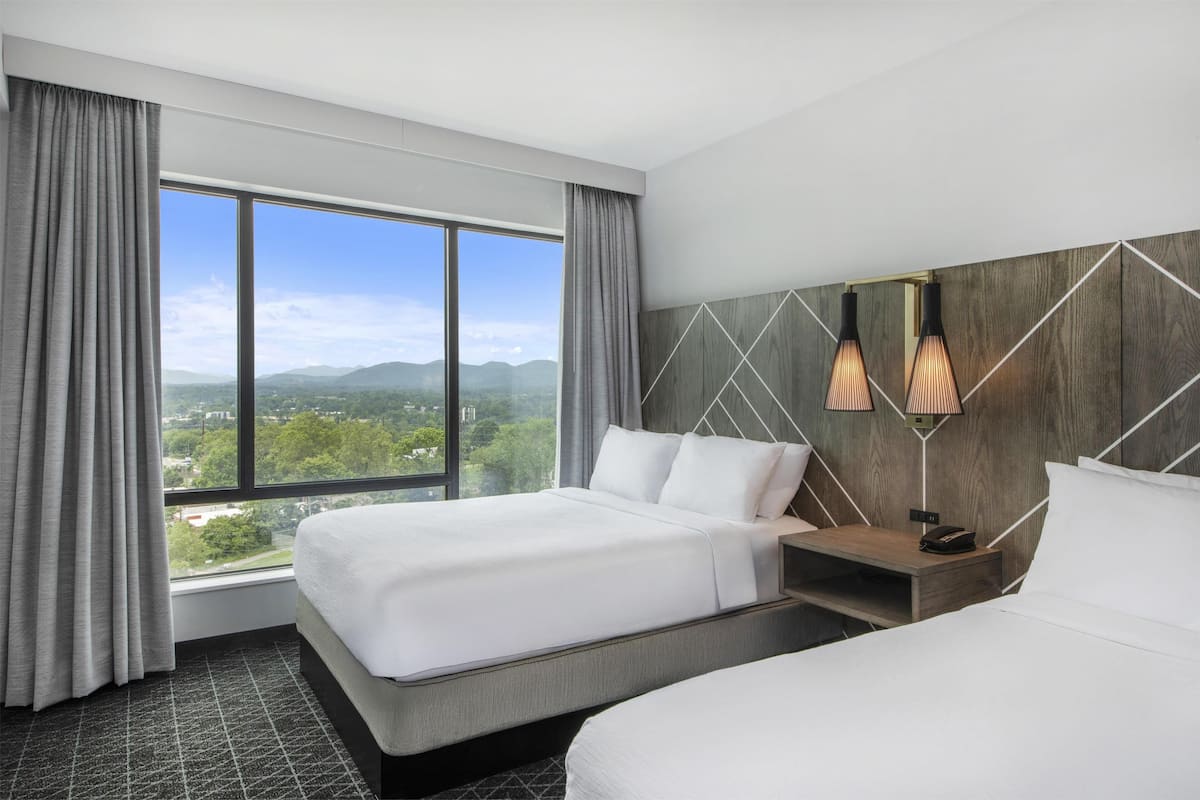 A hotel room with two beds, a large window with a view of mountains, and modern wall-mounted lighting.
