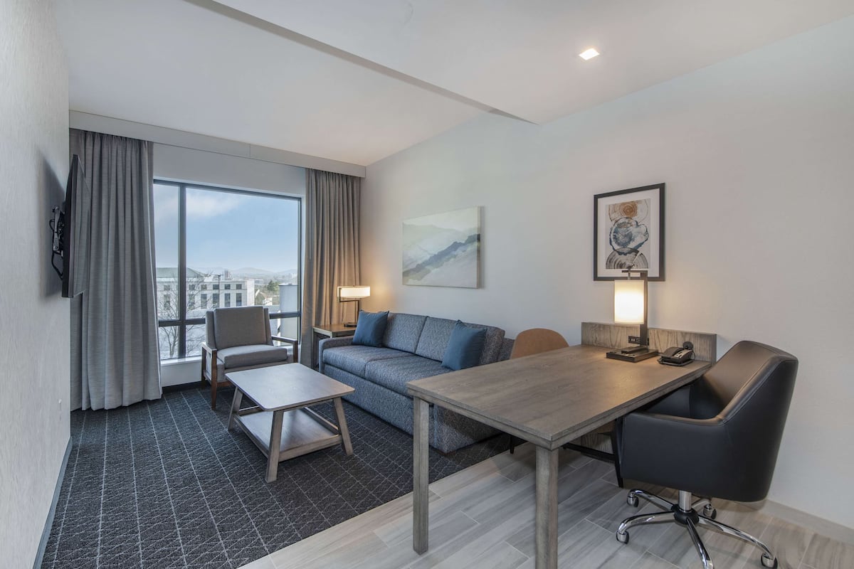 A modern hotel room with a large window, a sofa, a dining table, and a desk with a chair.