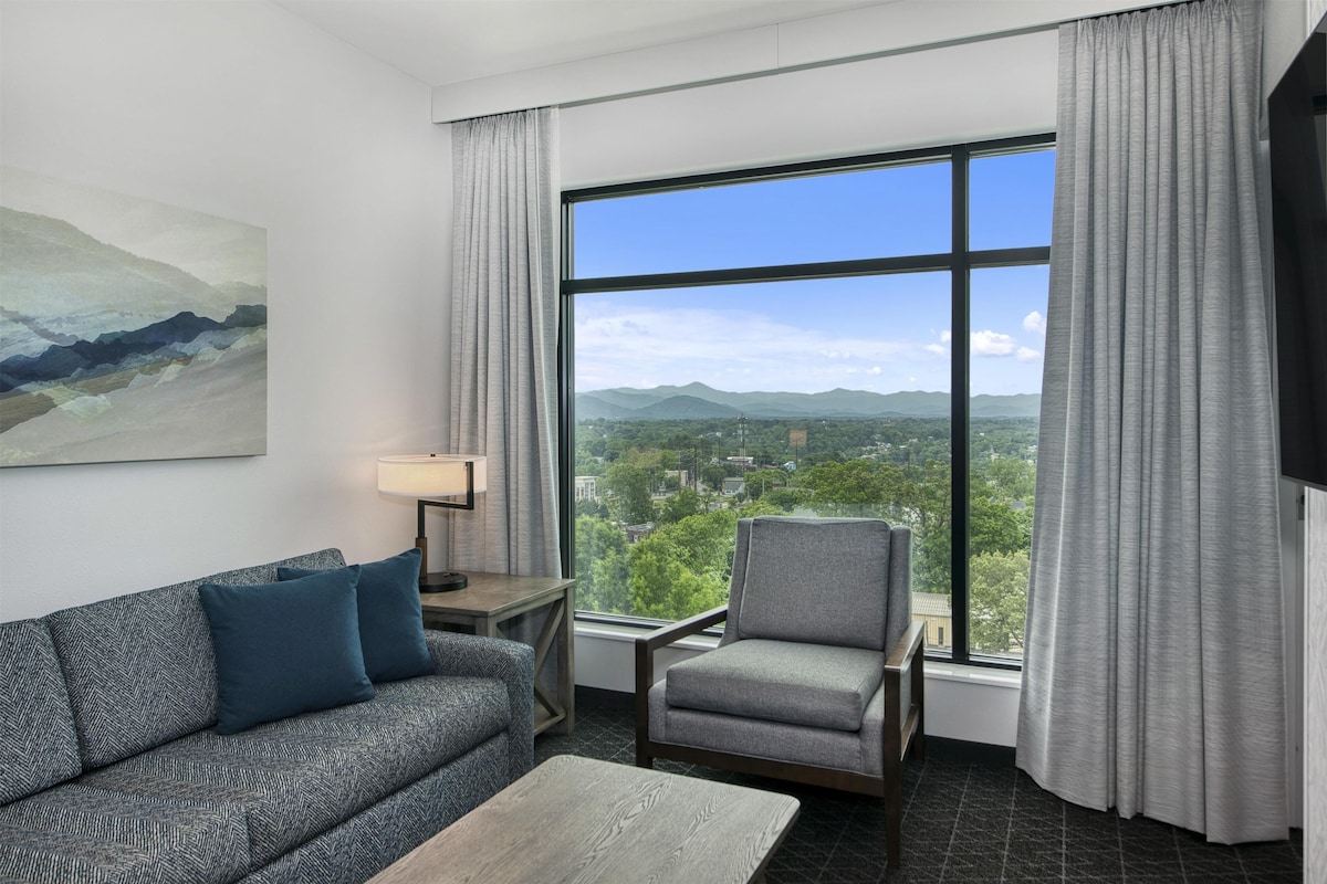 A modern hotel room with a large window offering a cityscape view, a sofa, a chair, and a small table.
