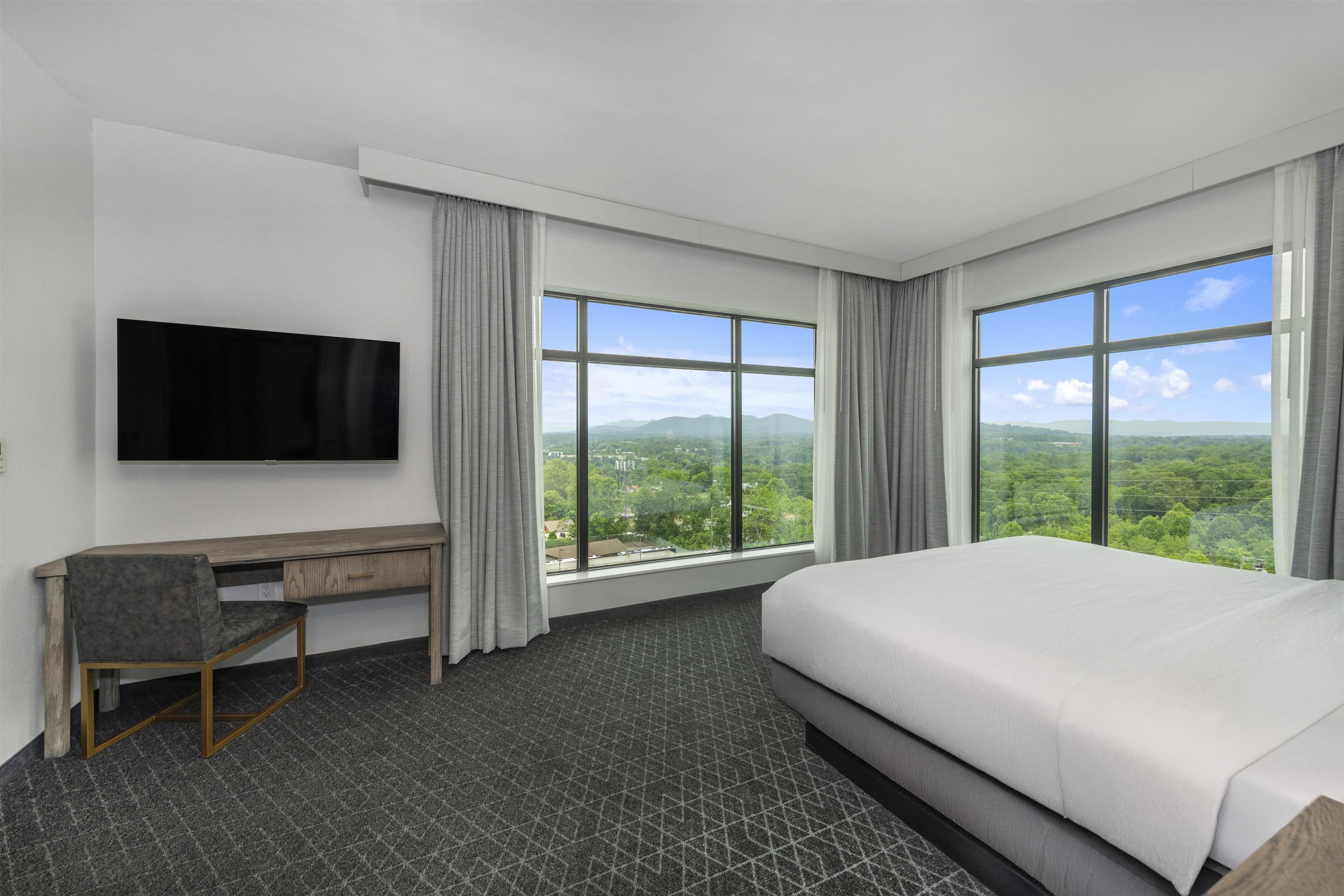 A hotel room with a large bed, a desk, a flat-screen TV, and large windows offering a view of the outdoors.