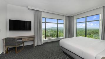 A hotel room with a large bed, a desk, a flat-screen TV, and large windows offering a view of the outdoors.