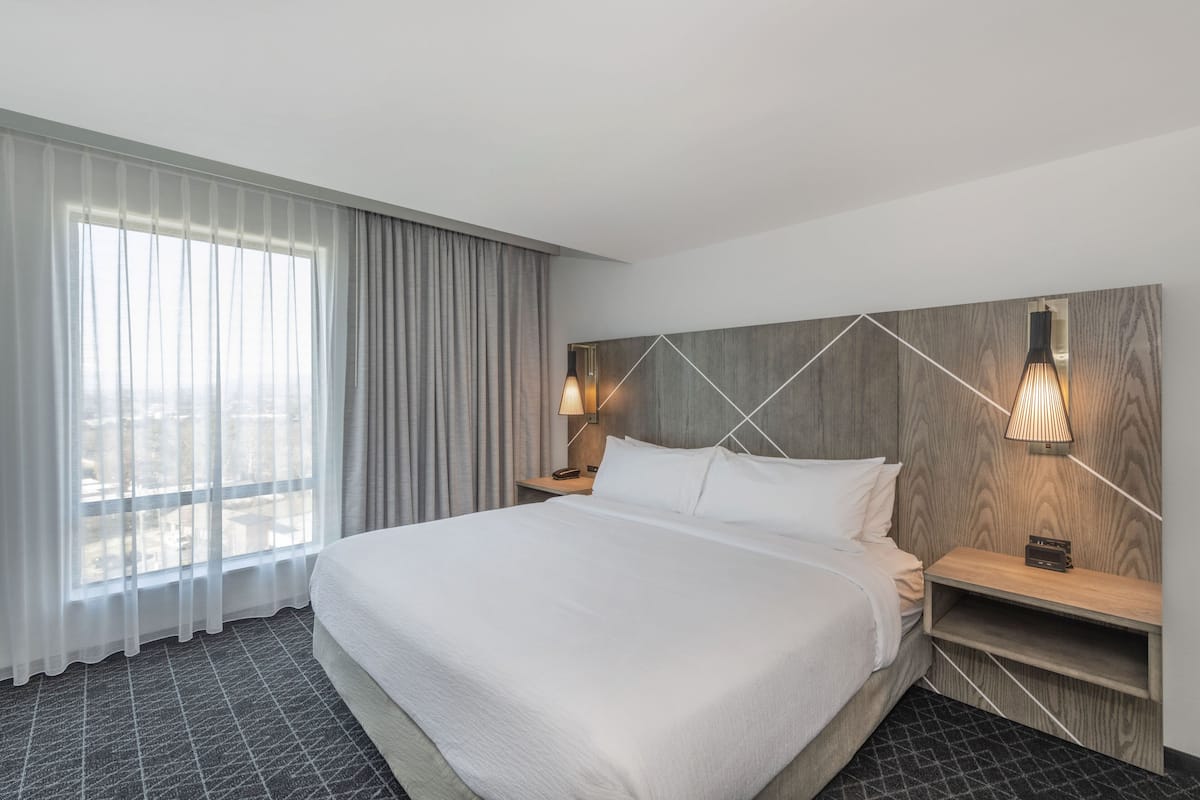 A hotel room with a large bed, a wooden headboard, two bedside lamps, a nightstand, and a view of the city through sheer curtains.