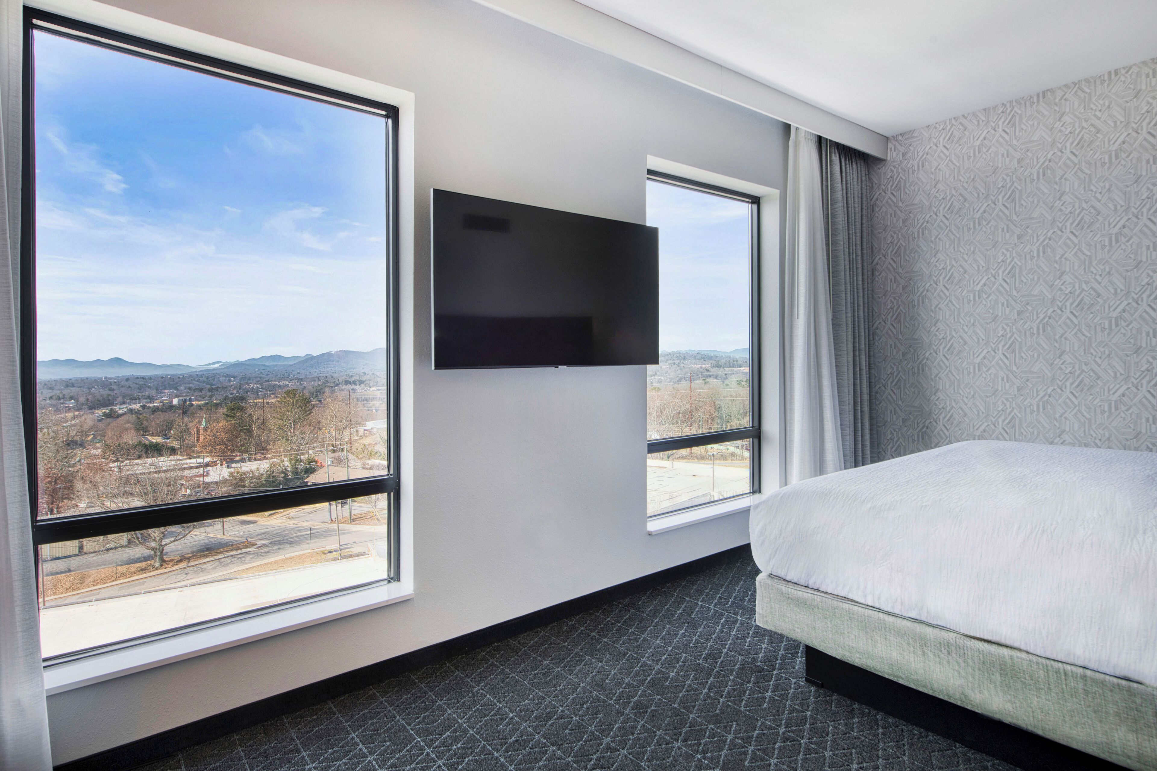 A hotel room with a large bed, a flat-screen TV mounted on the wall, and a view of a cityscape through large windows.