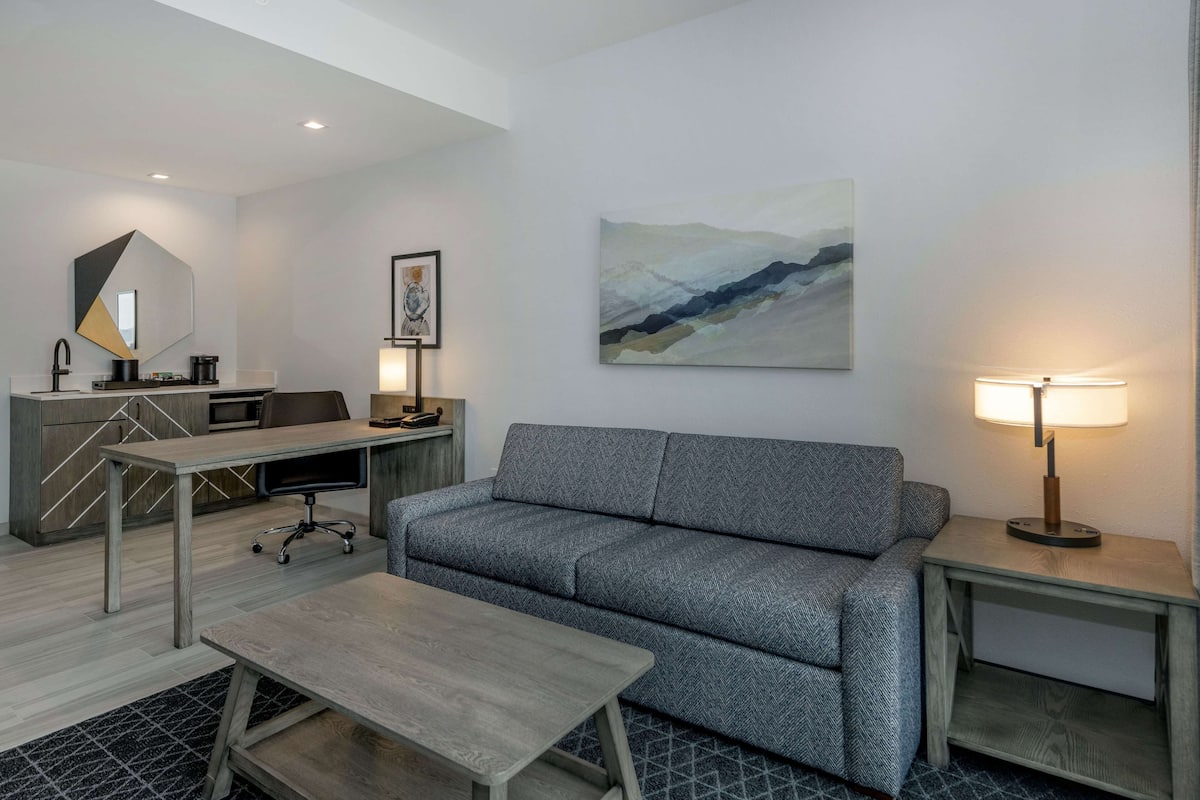 A modern hotel room with a grey sofa, a wooden coffee table, a desk with a chair, a lamp, and a painting on the wall.