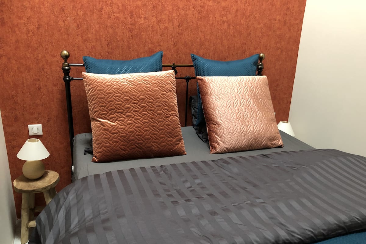 A bed with a dark grey bedspread, two patterned pillows, a wooden nightstand with a lamp, and a red textured wall.