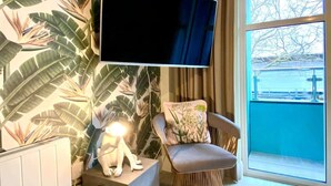A room with a large flat-screen TV mounted on the wall, a chair with a floral cushion, and a window with a view of a balcony.