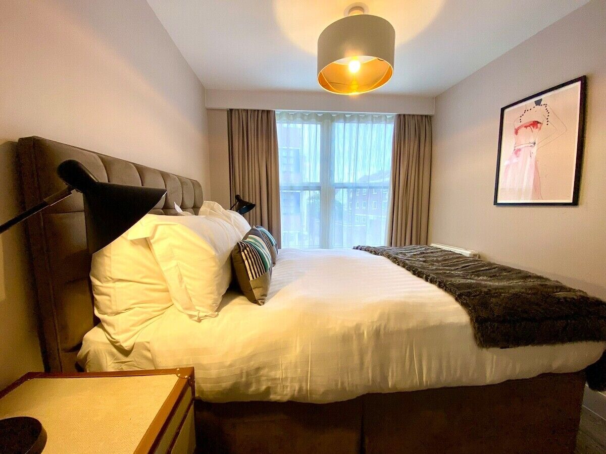 Une chambre d’hôtel avec un lit, une table de chevet, une lampe, une fenêtre avec des rideaux et un tableau encadré au mur.