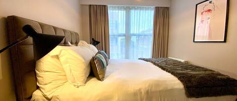 Habitación de hotel con cama, mesita de noche, lámpara, ventana con cortinas y un cuadro en la pared.