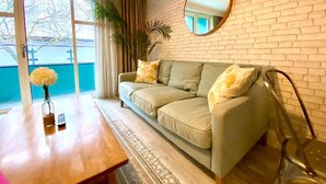 A living room with a sofa, a coffee table with a vase and remote, a mirror on the wall, and a view of the outdoors.