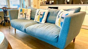 A blue sofa with patterned cushions in a room with wooden flooring and a kitchen area in the background.