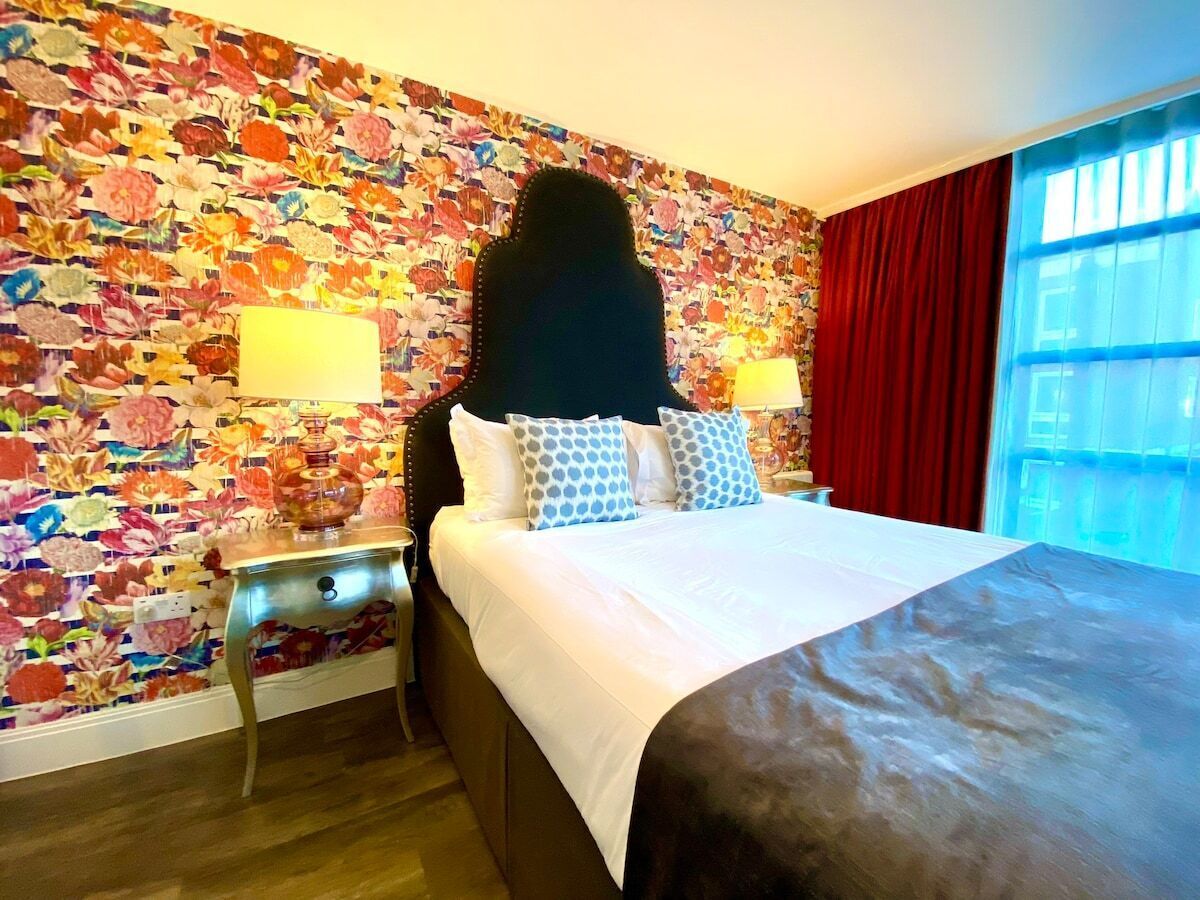 Une chambre d’hôtel avec un papier peint à motifs floraux, un lit avec du linge de maison blanc et des oreillers à motifs, une table de chevet avec une lampe et une fenêtre avec des rideaux rouges.