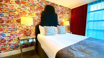 Une chambre d’hôtel avec un papier peint à motifs floraux, un lit avec du linge de maison blanc et des oreillers à motifs, une table de chevet avec une lampe et une fenêtre avec des rideaux rouges.