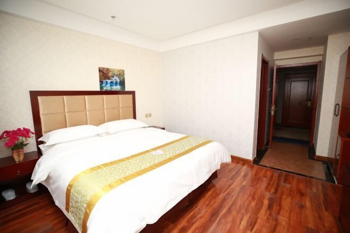 A hotel room with a large bed, wooden flooring, a painting on the wall, and a door leading to another room.