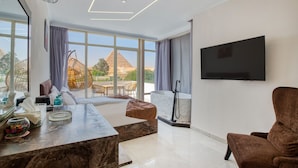 A modern hotel room with a large window, a flat-screen TV, a sofa, a coffee table, and a view of a pyramid. - Shadow Pyramids View INN (Giza)