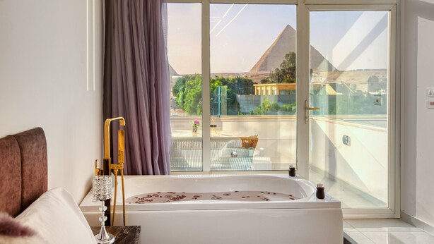 A bedroom with a large window, a bathtub, a bed, and a view of a pyramid. - Shadow Pyramids View INN (Giza)