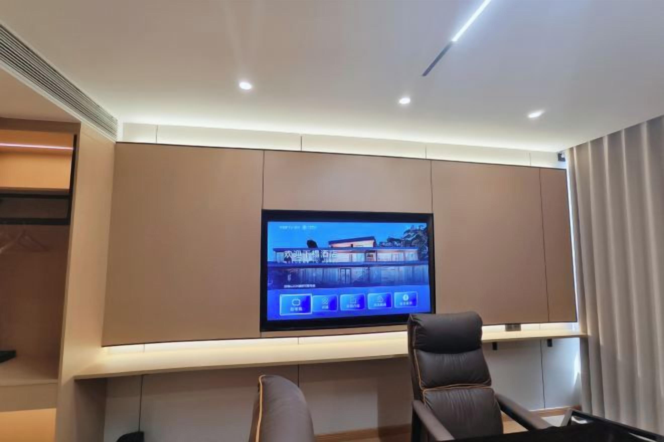 Een moderne hotelkamer met een grote flatscreen-televisie aan de muur, een comfortabele zithoek met een bank en fauteuils, en een houten bijzettafeltje.