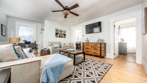 A modern living room with a sofa, coffee table, and TV. There is a ceiling fan, a patterned rug, and a wooden cabinet. - Steps to Harbor & Dining! Updated Home Slps 12 (Hyannis)