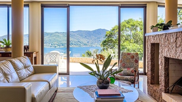 A spacious living room with a large window offering a view of the sea and mountains. There is a sofa, a coffee table with books, and a patterned armchair. - Refúgio Mirante da Lagoa - Floripa (Florianópolis)