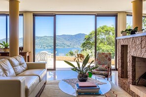 A spacious living room with a large window offering a view of the sea and mountains. There is a sofa, a coffee table with books, and a patterned armchair. - Refúgio Mirante da Lagoa - Floripa (Florianópolis)