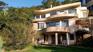 A modern two-story house with a curved balcony, surrounded by lush greenery. - Refúgio Mirante da Lagoa - Floripa (Florianópolis)