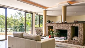 A modern living room with a beige sofa, a fireplace, and large windows offering a view of greenery outside. - Refúgio Mirante da Lagoa - Floripa (Florianópolis)