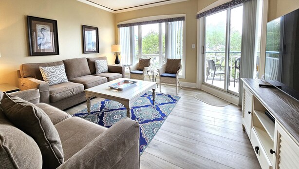 A living room with a sofa, armchairs, a coffee table, and a TV. There are large windows with curtains and a view of greenery outside. - Amazing Villa w/ Beautiful Views from Balcony! (Hilton Head Island)