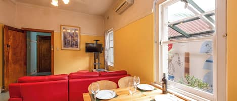A dining area with a wooden table set for two, a red sofa, and a television on the wall.
