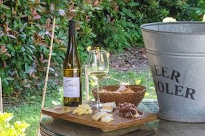 Una botella de vino, una copa, queso, galletas y un enfriador de cerveza sobre una mesa al aire libre.