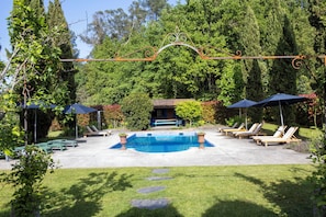 Piscina con sillas de descanso y sombrillas, rodeada de árboles y un pequeño edificio.