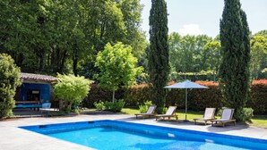 A rectangular swimming pool with clear blue water, surrounded by a concrete deck. There are two lounge chairs and a small umbrella providing shade.