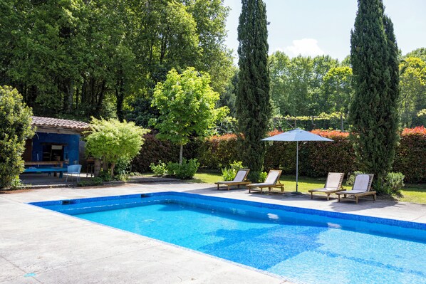A rectangular swimming pool with clear blue water, surrounded by a concrete deck. There are two lounge chairs and a small umbrella providing shade.