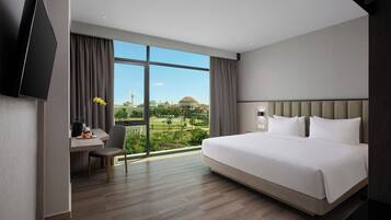 A modern hotel room with a large bed, a desk with a chair, a flat-screen TV, and a view of a golf course and buildings.
