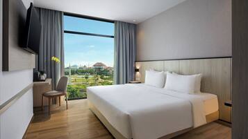 A modern hotel room with a large bed, a flat-screen TV mounted on the wall, a desk with a chair, and a view of a golf course and buildings through large windows.