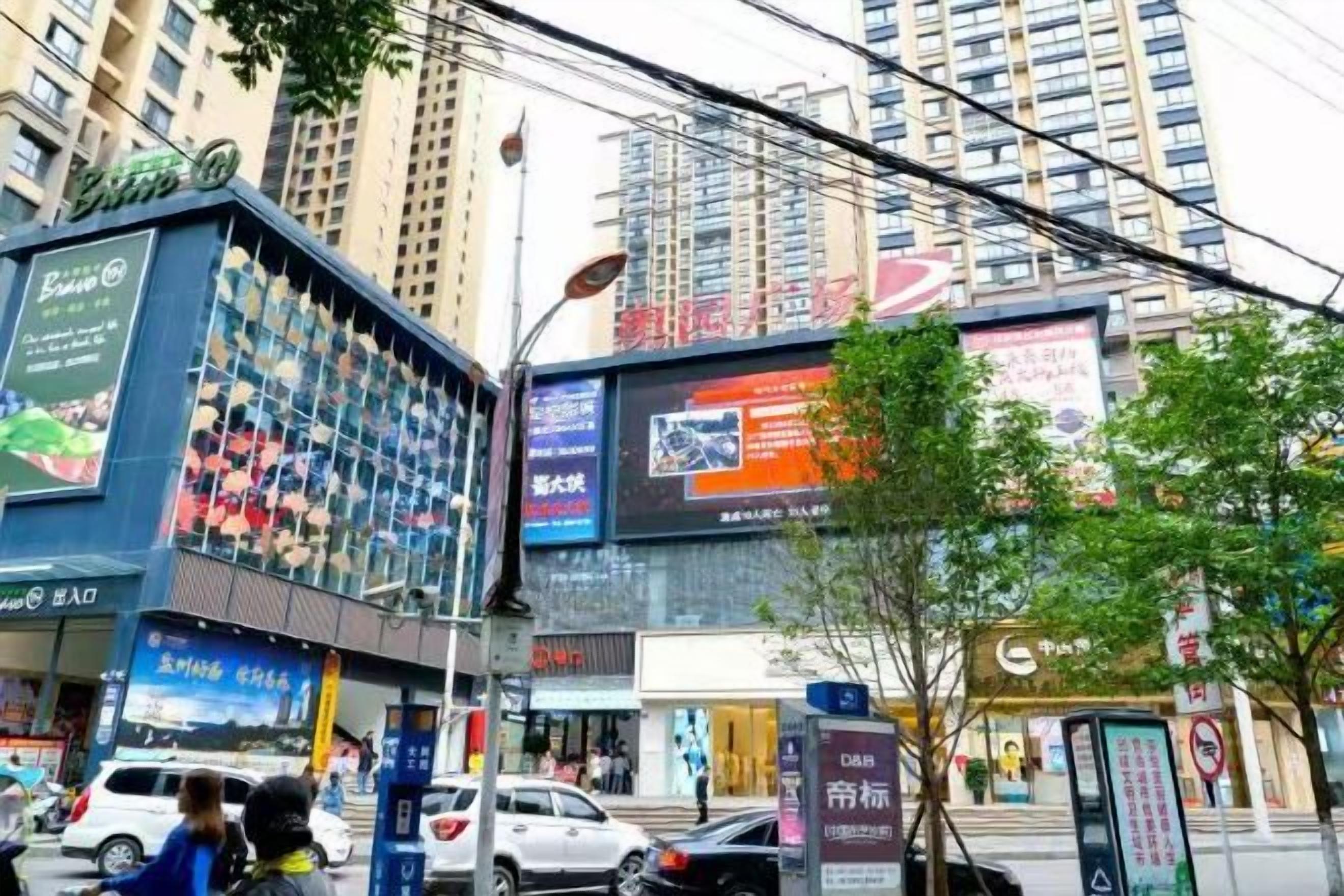 A street view with a building featuring a large billboard, storefronts, and a tree.