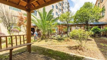 A covered outdoor area with a wooden deck, surrounded by greenery and buildings.