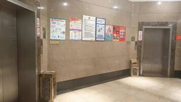 Zona de ascensores con suelo de baldosas, una pared con carteles informativos y una puerta de ascensor cerrada.
