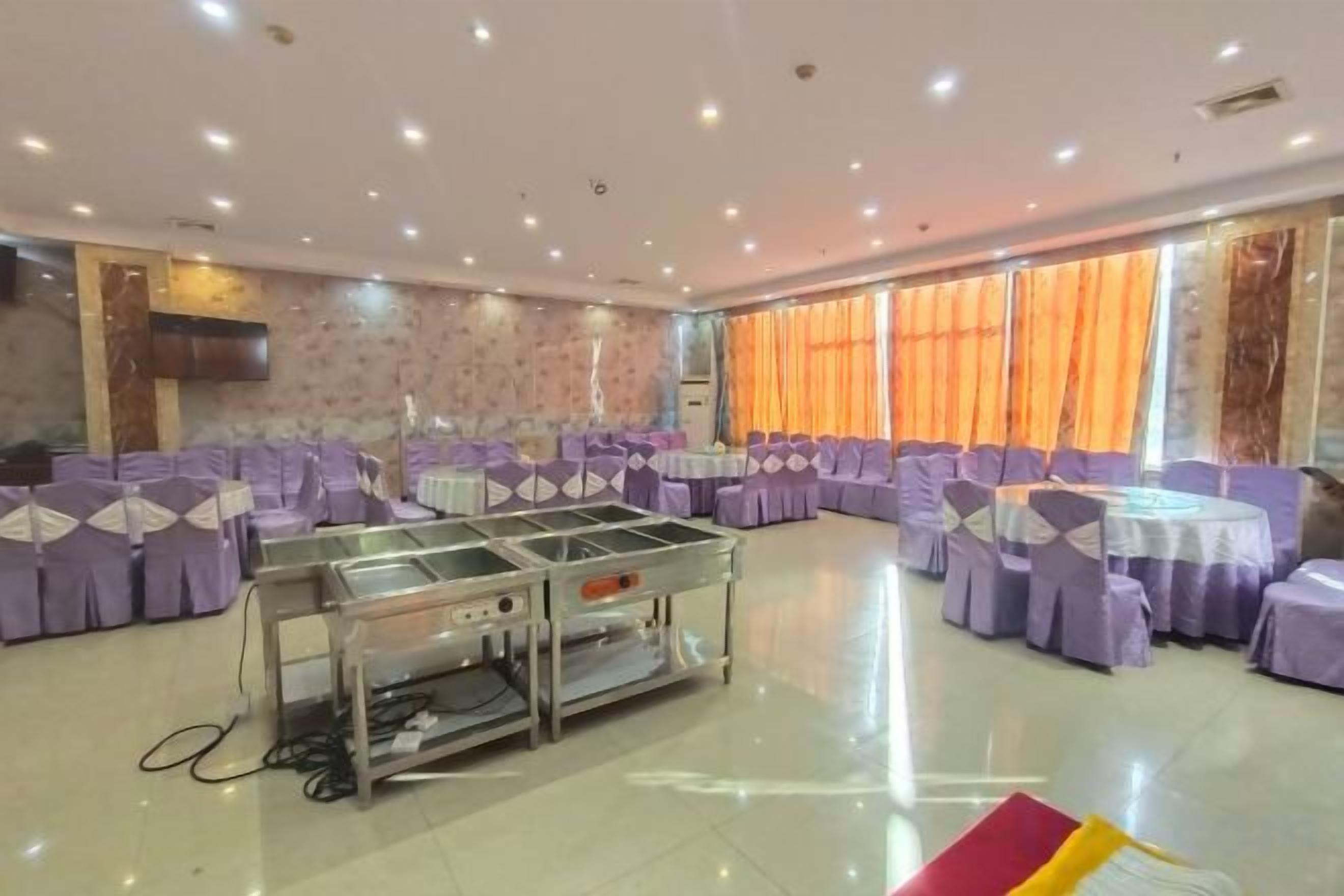 A banquet hall with round tables and chairs, a buffet station, and a large window with curtains.