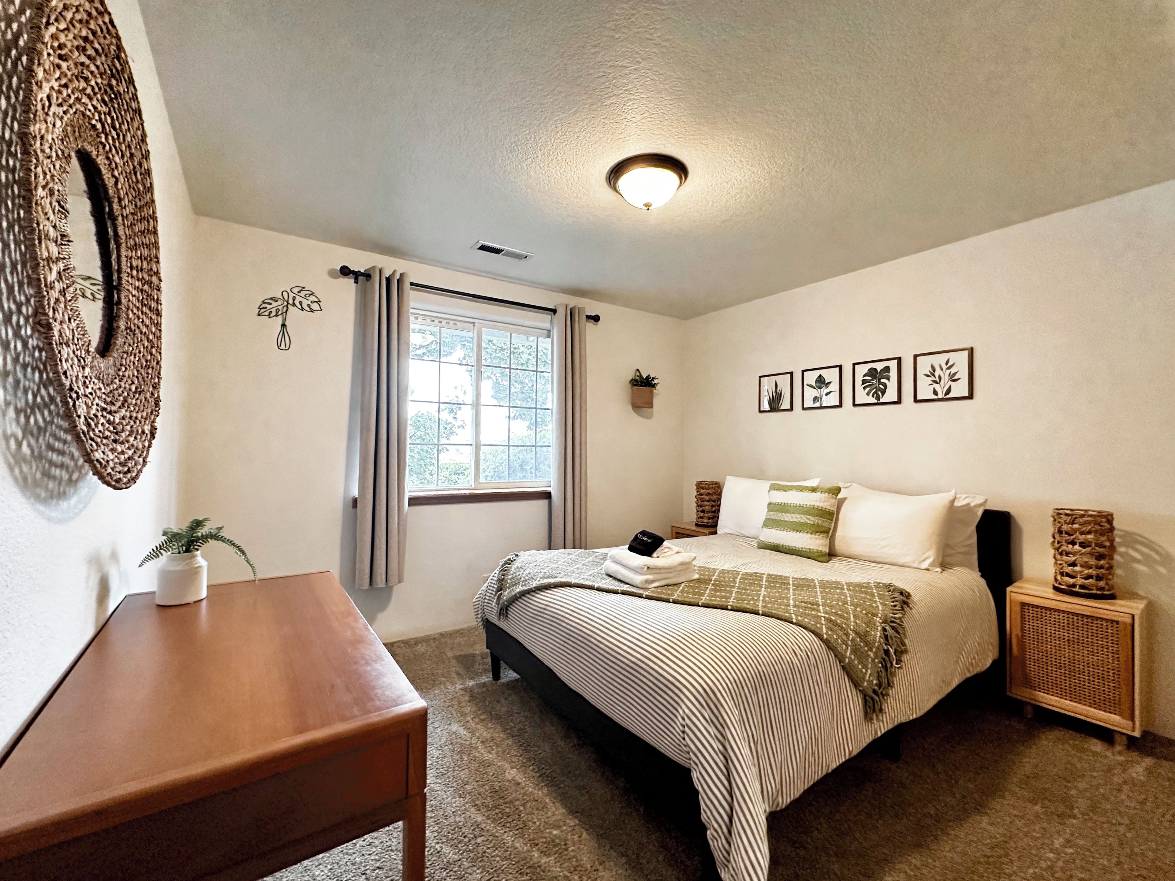 A bedroom with a bed, bedside tables, a window with curtains, a wall-mounted mirror, and framed pictures.