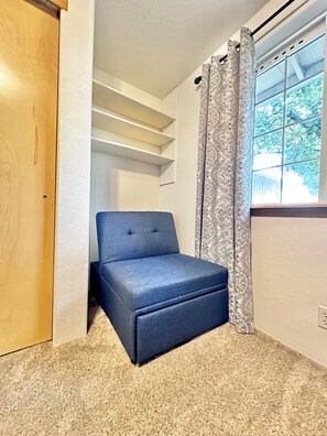Ein kleines Zimmer mit einem blauen Sofa, einer Holztür, einem Fenster mit gemusterten Vorhängen und einem Teppichboden.