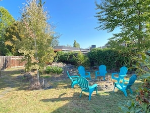 Ein Garten mit Holzpforte, einem Baum und vier blauen Adirondack-Stühlen, die um eine kleine Feuerstelle angeordnet sind.