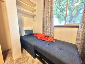 Una habitación pequeña y funcional con una cama individual, una ventana con cortinas y una estantería empotrada.
