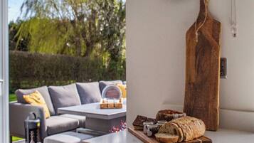 Un espace extérieur aménagé avec une table et des chaises, une planche à découper en bois sur laquelle sont disposés du pain et des condiments, et un porte-couteaux fixé au mur.