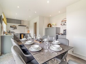 A modern dining area with a long table set for six, a kitchen in the background, and a white radiator.