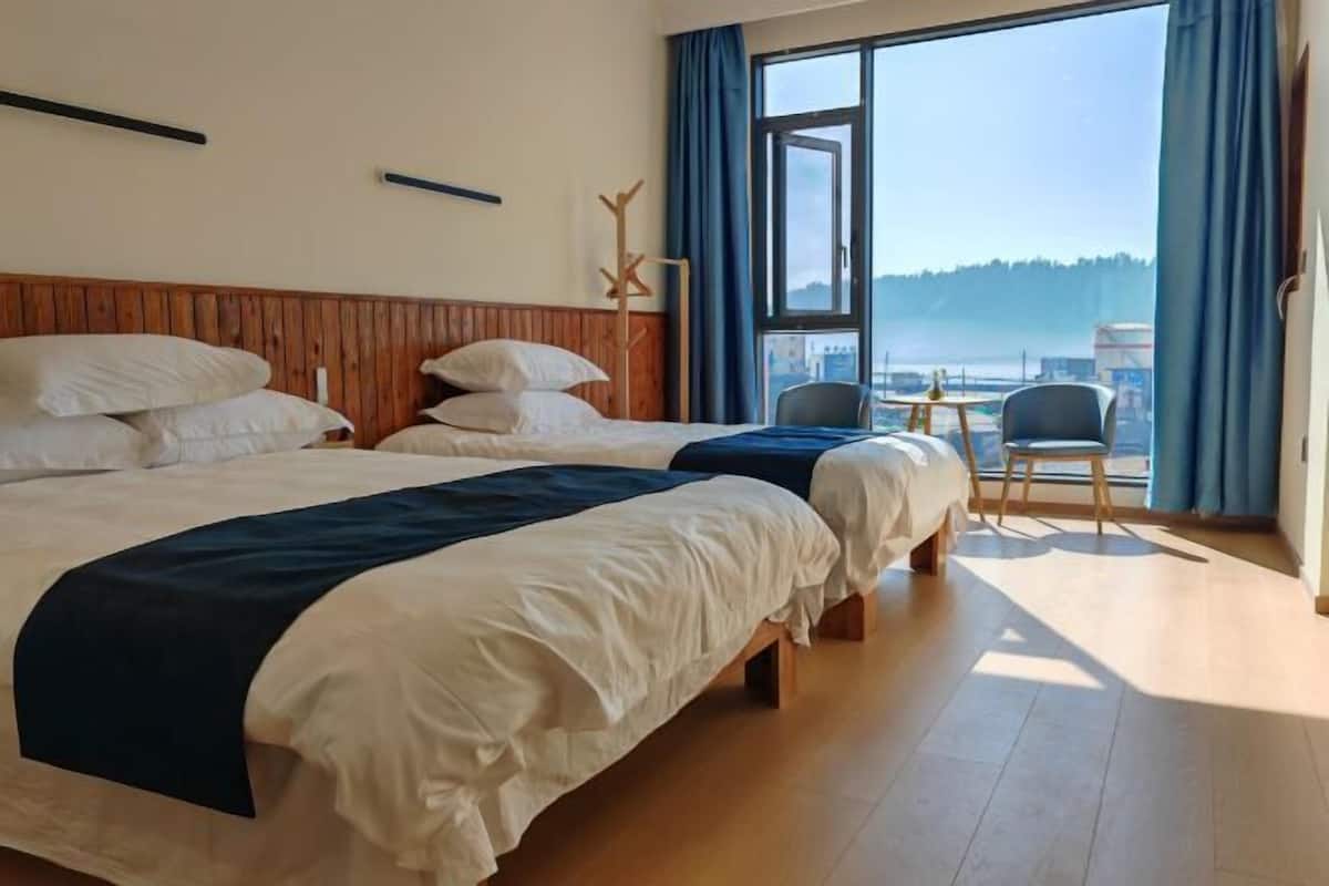 A hotel room with two beds, a small table, chairs, a large window with a view, and a wooden headboard.