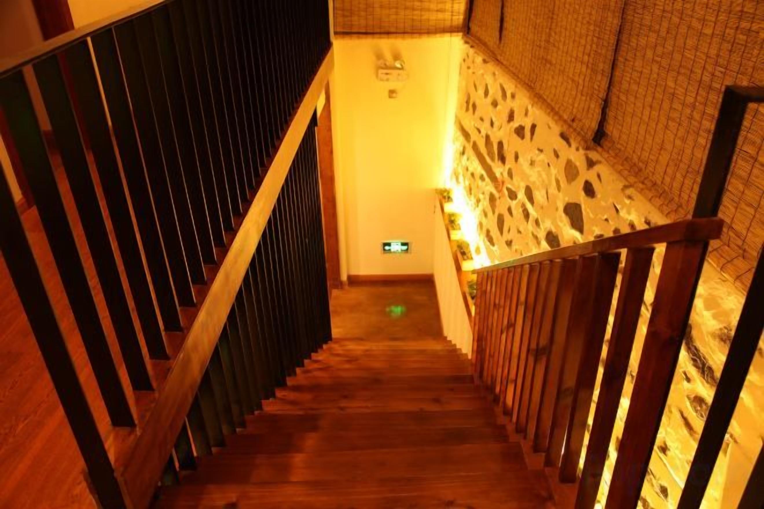 A wooden staircase with a metal railing leading to a room with a patterned wall and a green exit sign.