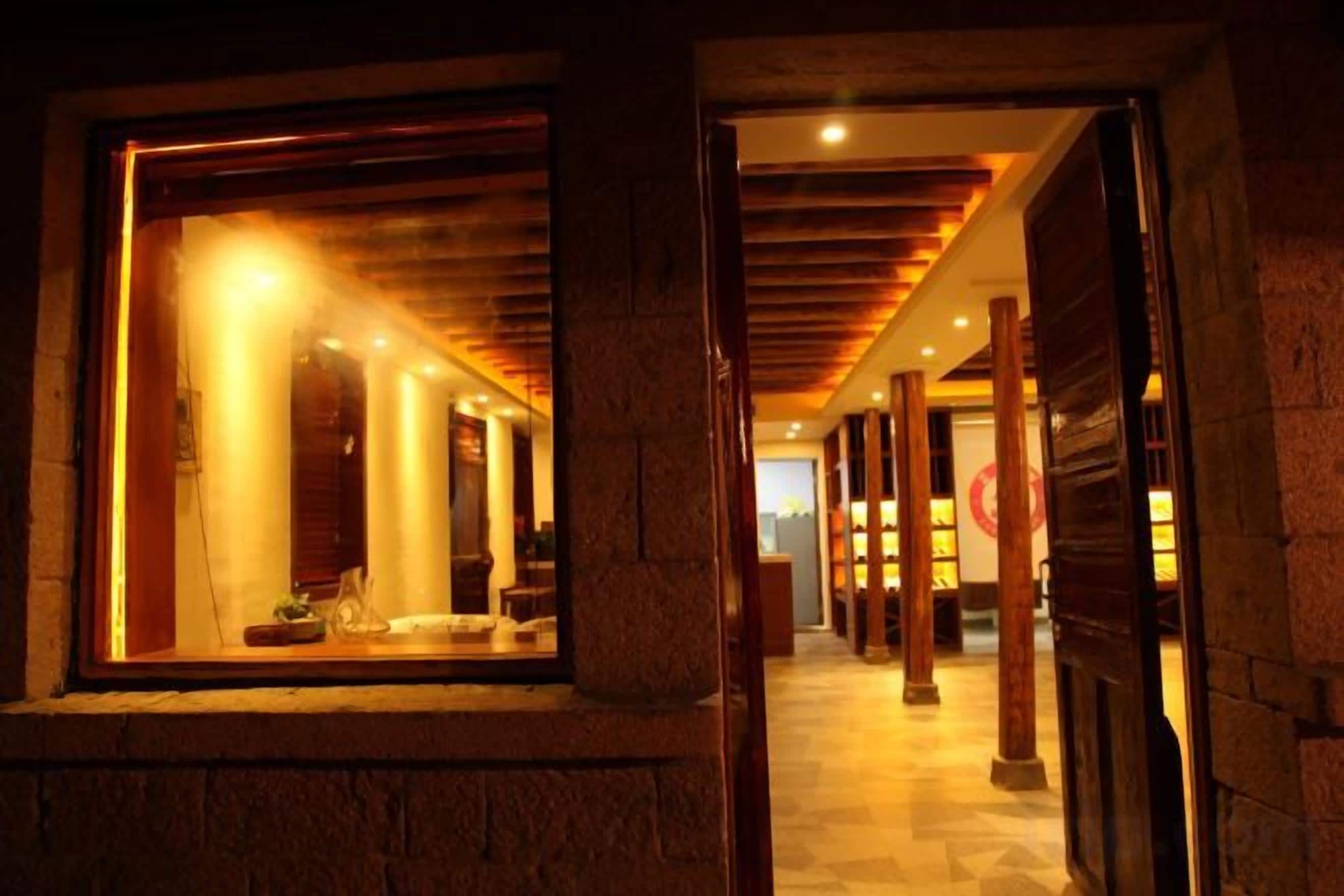 A corridor with wooden beams and a stone wall, leading to a room with a large window and a bed.