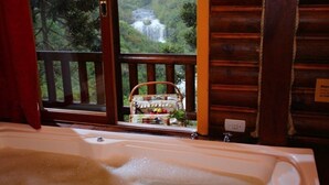 A bathroom with a bathtub, a window with a view of a waterfall, and a basket of toiletries on the windowsill. - Cabanas Cambuim - Urubici - SC (Urubici)