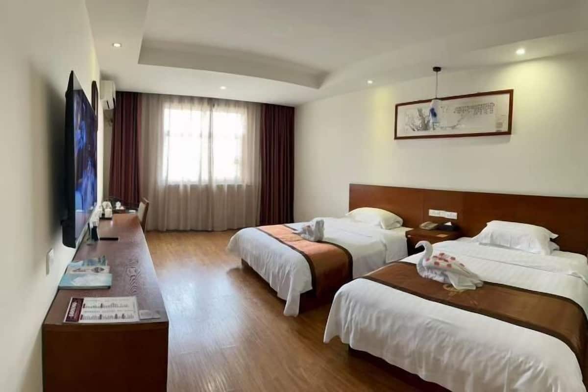 A hotel room with two beds, a desk with a flat-screen TV, a large window with curtains, and a framed picture on the wall.