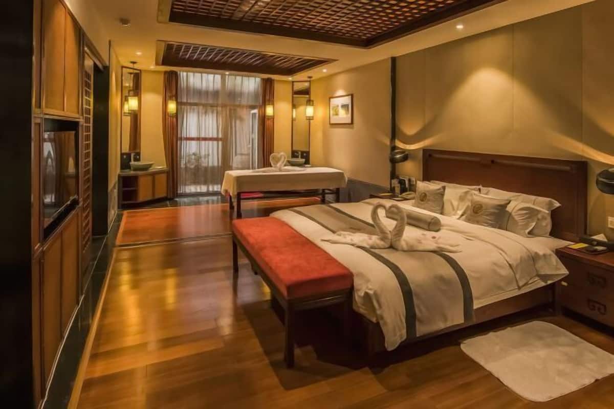 A hotel room with a wooden floor, a bed with swan-shaped pillows, a bench, a small table, and a mirror.